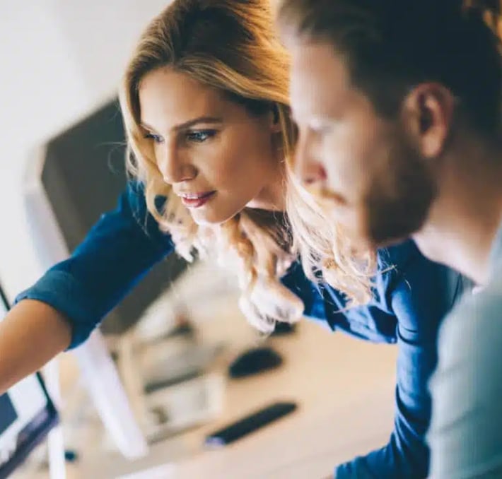 Two people work together at a computer desk, with the woman pointing at the screen while the man looks on attentively as they explore the buyer’s journey with CPQ.