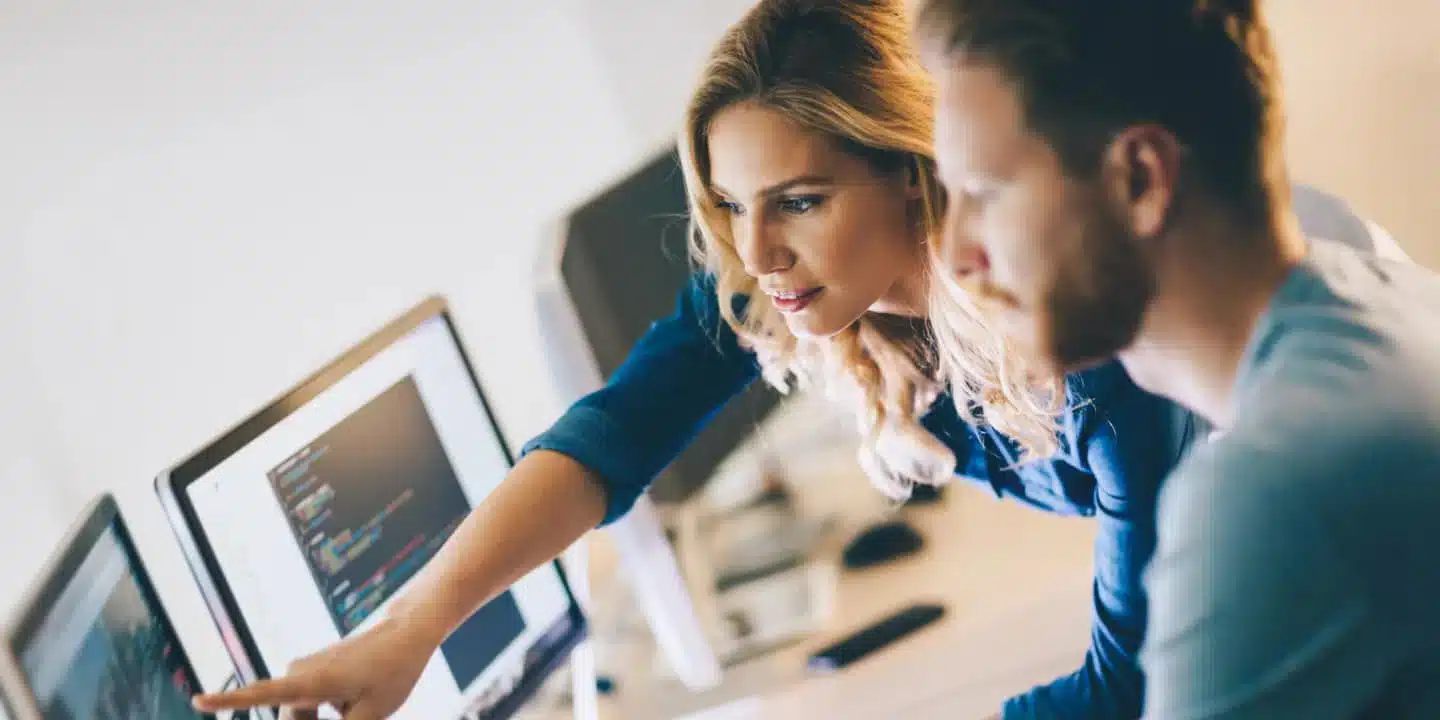 A woman and a man look at computer monitors displaying code while the woman points at the screen.