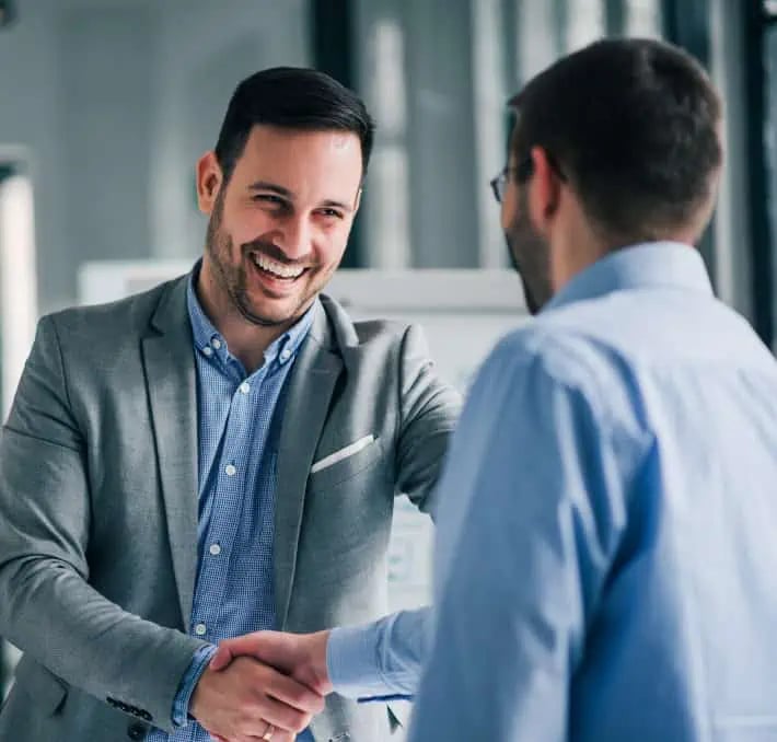 Two men in business attire shake hands and smile in an office setting.