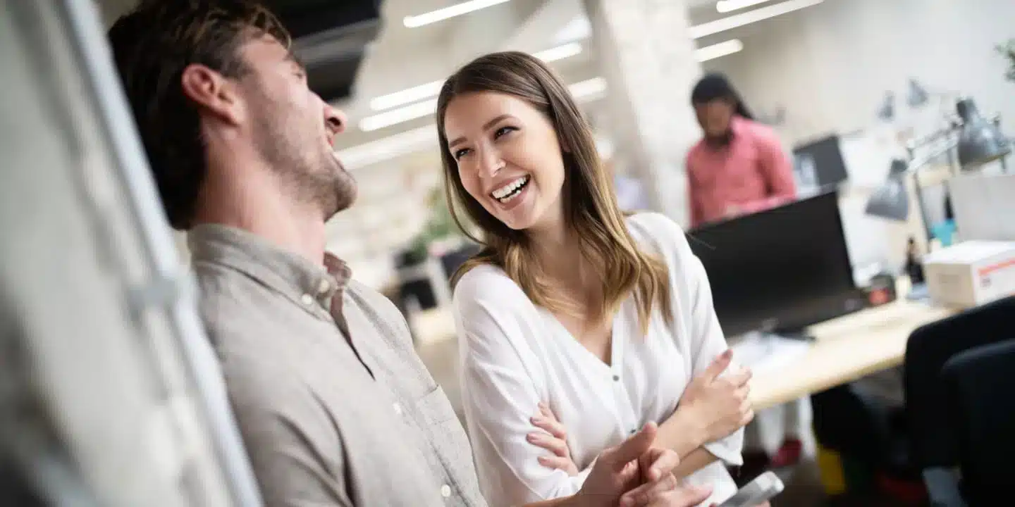 Two colleagues stand and chat, smiling and laughing in a modern office with desks, computers, and another person working in the background.