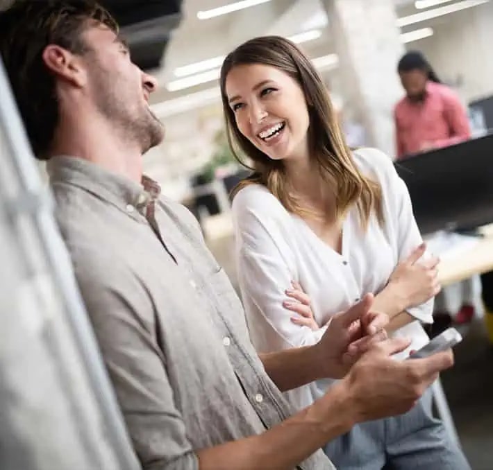 Two people standing in an office, smiling and talking casually; one holds a smartphone while others work at desks in the background.