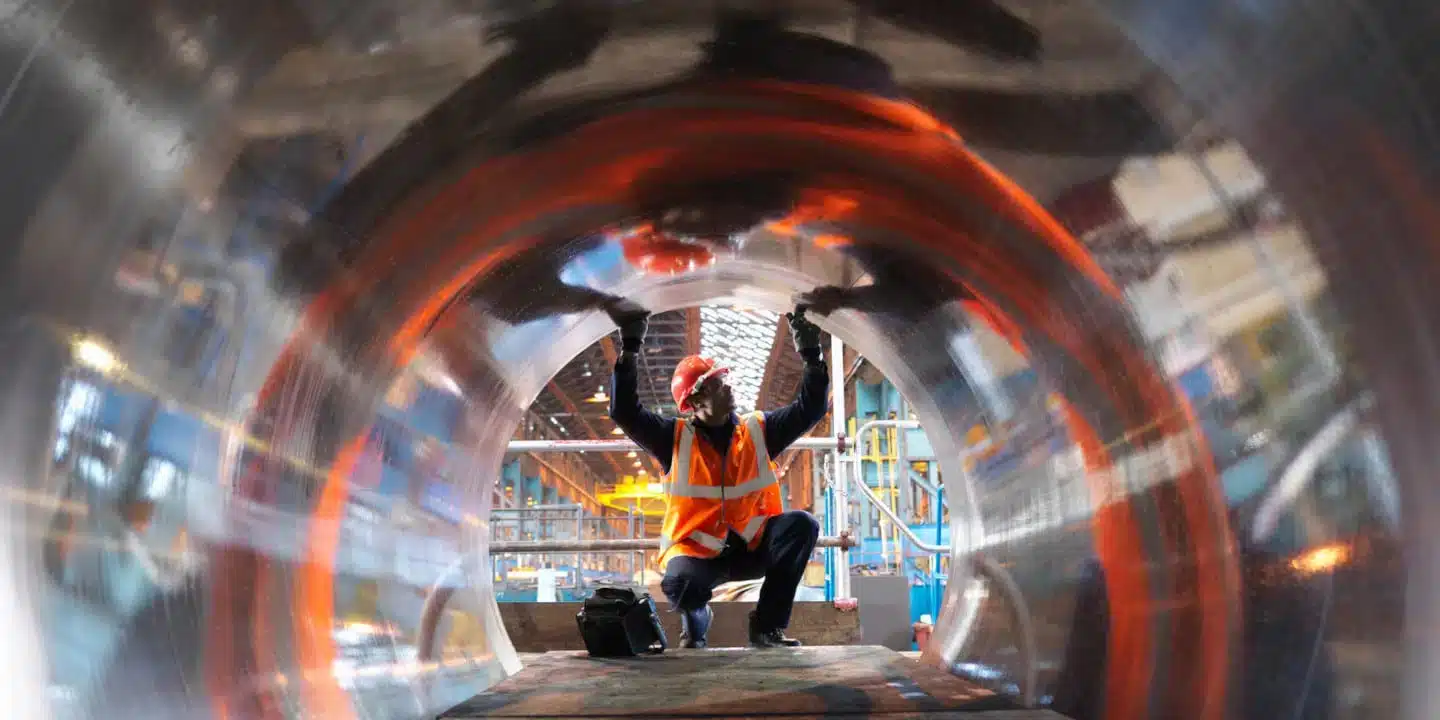 Worker in an orange vest and hard hat inspects the inside of a large cylindrical metal pipe at an industrial site.