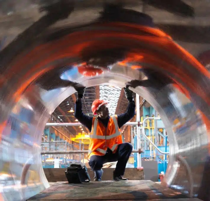 A worker in a hard hat and safety vest inspects the interior of a large metal cylinder in an industrial facility.