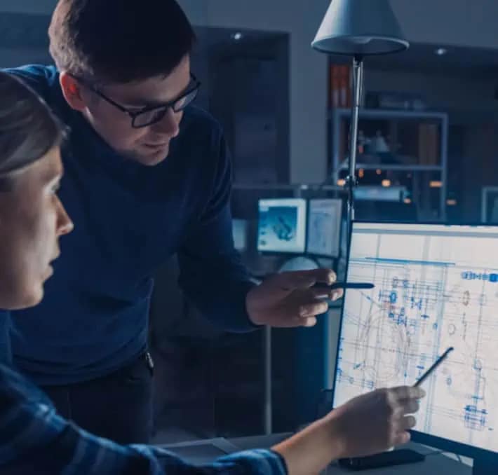 Two people in an office discuss a technical diagram displayed on a computer monitor, with one person pointing at the screen.