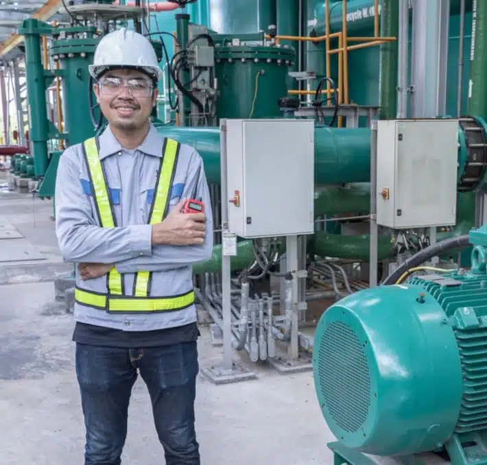 A worker wearing a safety helmet, glasses, and a reflective vest stands with arms crossed in an industrial facility with green machinery and control panels.