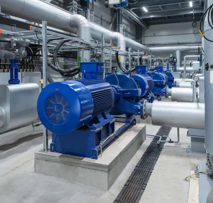 Rows of large industrial electric motors and pumps in a modern facility, connected by metal pipes amid various equipment and overhead cables.