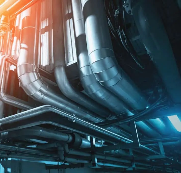 Large metallic pipes and ducts run along a ceiling in an industrial or mechanical facility, illuminated by both warm and cool lighting.