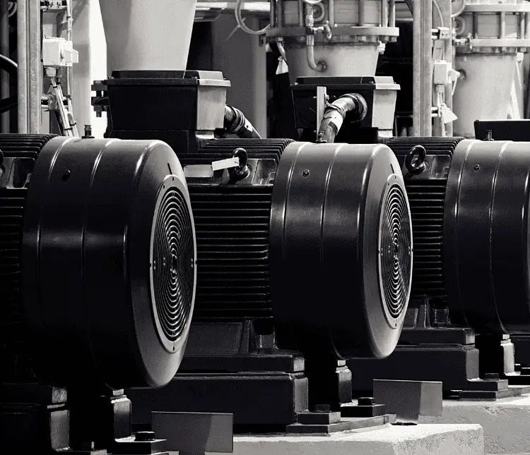 Row of large industrial electric motors with visible vents and wiring, installed on a concrete platform in a factory setting.