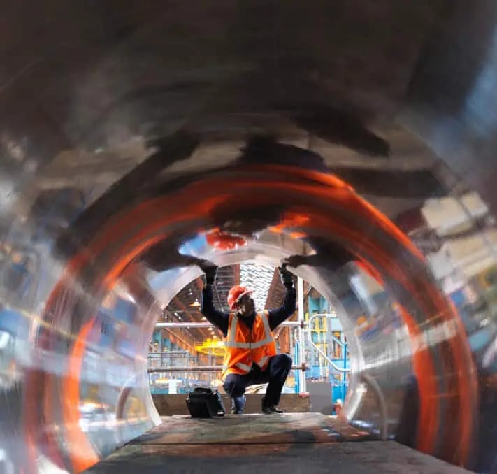 A worker in a safety vest and helmet inspects the interior of a large metal pipe at an industrial facility.