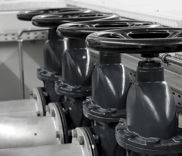Close-up view of four large industrial valve wheels in a row, attached to dark metal piping, with a background of equipment and metal framework.